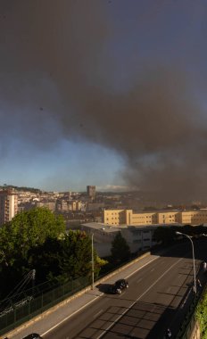 Şehir üzerinde yoğun bir kara duman sütunu yükseliyor, binalar, önünde arabalar ve ağaçlar olan bir yol, ve temiz hava ile duman arasında net bir ayrım yapan mavi bir gökyüzü..