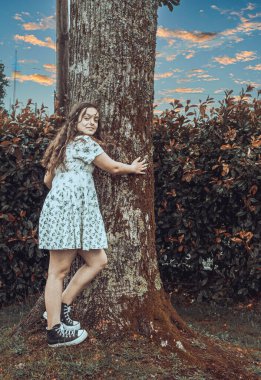 Kıvırcık saçlı ve açık renk elbiseli genç bir kadın bir ağacın gövdesine sarılıyor. Arka plan yeşilliği ve gün batımında bulutlu gökyüzünü gösteriyor, doğayla bir bağlantı kuruyor..