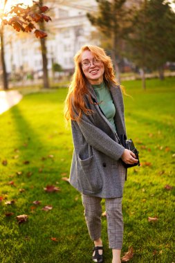 Redhead smiling pretty woman walking in park on sunny autumn day. Elegant female poses in stylish authentic outfit has fun laughing on green lawn with vivid foliage, leaves. Girl enjoys a fall season.