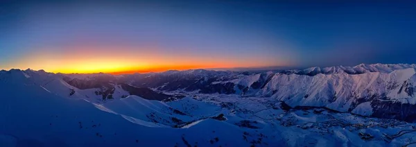 Wide aerial panorama of snowy mountain ridge on winter sunrise. Stunning mountains range covered with snow powder on ski resort at sunset. Caucasus mountain peaks skyline in a dusk golden hour.