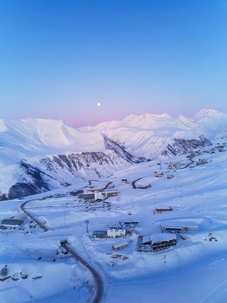 Aerial of snowy mountain range on winter sunrise at ski resort. Moon above mountains valley and village with road curves at sunset. Caucasus peaks skyline in a pink twilight afterglow.