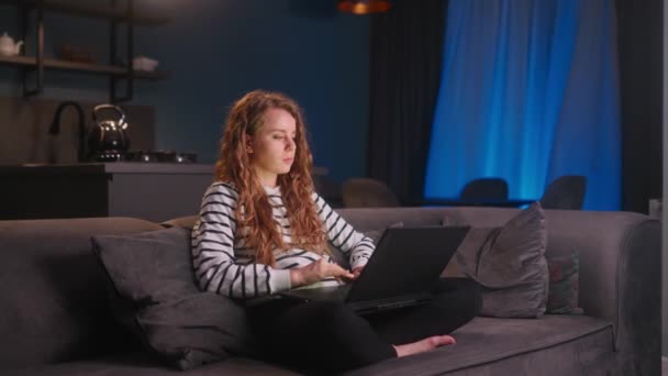 オンライン研究 宿題のためのコンピュータを使用して焦点を当てた女の子 自宅からノートパソコンを持って仕事をしている若いフリーランスの女性は 夜にリビングルームのソファに座っています リモートジョブプロジェクトのためのキーボードを入力する女性 — ストック動画