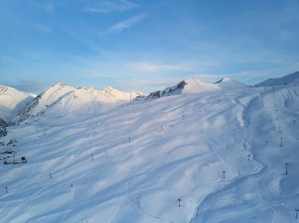 Aerial of snowy mountain slopes full of powder for freeride at ski resort on winter sunrise. Mountains range of backcountry covered with virgin snow. Caucasus peaks skyline with a twilight afterglow.