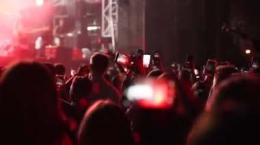 People waving hands silhouettes, taking photos or recording videos of live music concert with smartphones at open air festival. Bright colorful stage lighting. Fans at a favourite band performance.