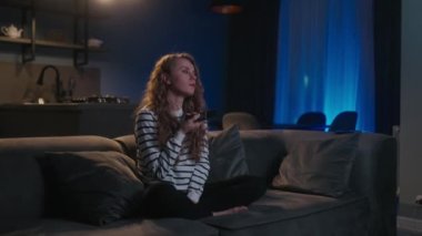 Şirin sarışın kıvırcık kadın televizyonu açıyor, akıllı TV uygulamalarında geziniyor ve evdeki oturma odasında kanepede otururken kendini eğlendirmek için kanalları seçiyor. Sinematik çekim.