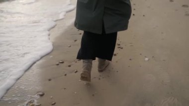 Kumlu deniz sahilinde kaçan mutlu kadının arka görüntüsü. Şık yeşil palto giyen, bej çizmeli ve dalgalar ve köpüklerle yıkanmış kıyı şeridi boyunca koşan bir kız. Yavaş çekimde rüya gibi sinematik bir çekim..