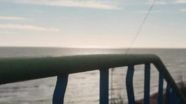 Kadın merdivenlerden aşağı iner, durur ve yıpranmış metal parmaklıkları tutarak deniz dalgalarına bakar. Yeşil ceketli bir kadın merdivenden iniyor, okyanus kıyısındaki parmaklıklara dokunuyor. Rüzgâr dalgalı saçlar savurur