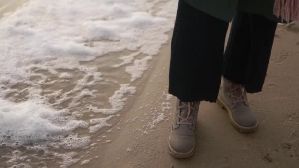 女人穿着靴子站在沙滩上被风吹来的泡沫中 动作缓慢 在狂风暴雨的日子里 雌性的脚穿着防水鞋 被海浪拍打在岸边的浮沫流过 低角度 — 图库视频影像
