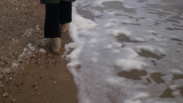 女人穿着靴子走在沙滩上被风吹来的泡沫中 动作缓慢 在多风的暴风雨天气里 雌性的脚在海滨被海浪拍打着 背景色 — 图库视频影像
