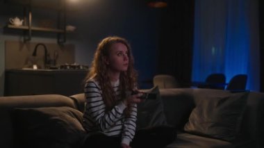 Şirin sarışın kıvırcık kadın televizyonu açıyor, akıllı TV uygulamalarında geziniyor ve evdeki oturma odasında kanepede otururken kendini eğlendirmek için kanalları seçiyor. Sinematik çekim.