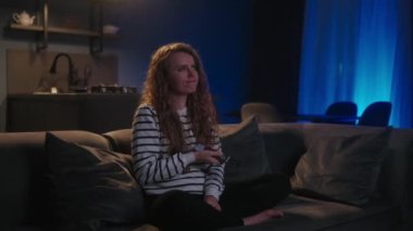 Şirin sarışın kıvırcık kadın televizyonu açıyor, akıllı TV uygulamalarında geziniyor ve evdeki oturma odasında kanepede otururken kendini eğlendirmek için kanalları seçiyor. Sinematik çekim.