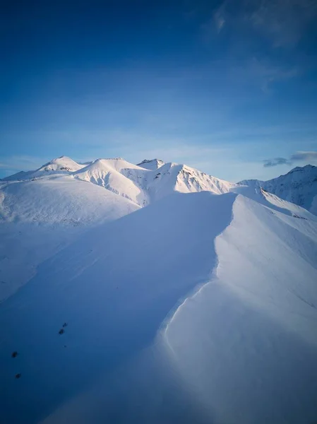 Aerial of snowy mountain slopes full of powder for freeride at ski resort on winter sunrise. Mountains range of backcountry covered with virgin snow. Caucasus peaks skyline with a twilight afterglow.