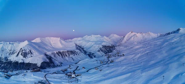 Wide aerial panorama of snowy mountain ridge on winter sunrise. Stunning mountains range covered with snow powder on ski resort at sunset. Moon above caucasus peaks skyline in a dusk at night. Gudauri