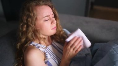 Soğuk algınlığı olan bir kadın evde ateşini kontrol etmek için battaniyenin altında öksürüyor ve burnunu siliyor. Beyaz kadın koronavirüsten muzdarip. 19 derece izole edilmiş. Termometreye bakıyor.