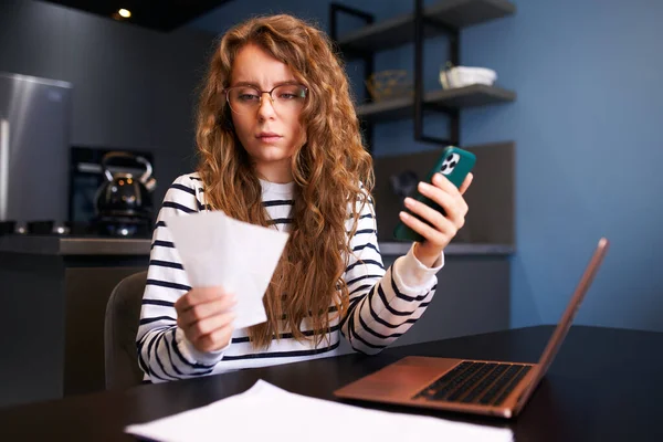 Focused curly woman calculates expenses on bills, increased utilities prices. Female counting income paychecks, debts, loans, reciepts to pay taxes. Inflation, financial planning, low paying capacity.