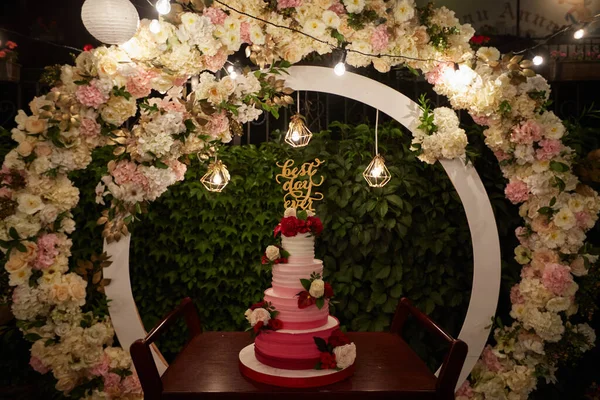 Wedding cake with best day ever sign on green garden decorated for wedding party at night. White, marsala creative decor with circle wedding arch decorated with roses and flowers, fairy lights.