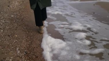Kadın deniz köpüğü boyunca botlarıyla yürüyor kumlu sahilde rüzgar tarafından ağır çekimde esiyor. Dişi ayaklar, fırtınalı bir günde okyanus dalgalarının kıyısında spume akıntısı parçalarıyla dolaşır. Arka plan