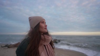 Pretty mixed ethnic woman looking at sunset with awe standing on ocean shore. Girl full of wonder watching, dreaming on sea coast. Waves breaking on stone groyne breakwater. Dreamy cinematic footage.