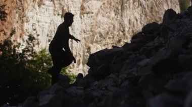 Silhouette of a hiker man climbing onto a rocky hill. A man hiking up a huge mountain field of rock. Silhouette of tourist climbing boulders in the mountains. Way to success. Struggle for achievements