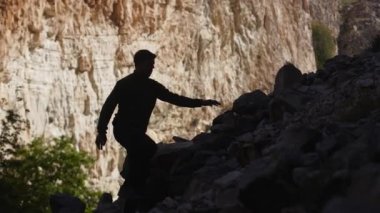 Silhouette of a hiker man climbing onto a rocky hill. A man hiking up a huge mountain field of rock. Silhouette of tourist climbing boulders in the mountains. Way to success. Struggle for achievements