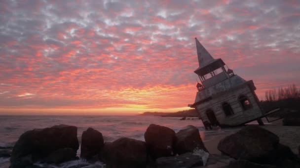 神秘的なヴィンテージは ピンクの夕日の海岸線で海の波に洗われた木の家やチャペルを傾け殺到した 日の出の防波堤の隣の砂浜の水の中に沈む壮大なレンガ造りの建物 — ストック動画