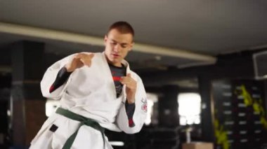 Martial arts man in kimonos practices kicks and punches. Male fighter wearing kimono training at gym. Kick boxer jumping and practicing kicking and punching actions indoors. People and sport concept