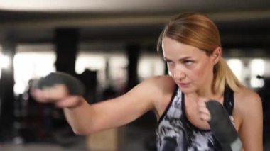 Female boxer is making punch moves with wrapped hands training at gym alone side view. Motivated female fighter with hands wrapped with bandage looking at camera and throwing punches jumping