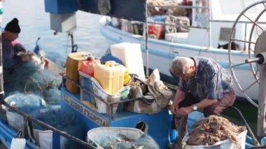 2019-05-01, Cyprus, Larnaka. Fishermen mending their nets on a fishing boat in harbor. Men mending fishing nets on moored boat in marina harbour. People fixing nets, one working on bow.