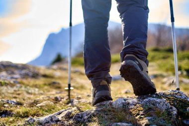 Legs of hiker in trekking boots walking in the mountains with Nordic walking poles closeup shot. Feet of walking tourist wearing trekking shoes with hiking sticks on rocky road captured from behind.