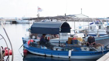 2019-05-01, Cyprus, Larnaka. Fishermen mending their nets on a fishing boat in harbor. Men mending fishing nets on moored boat in marina harbour. People fixing nets, one working on bow.