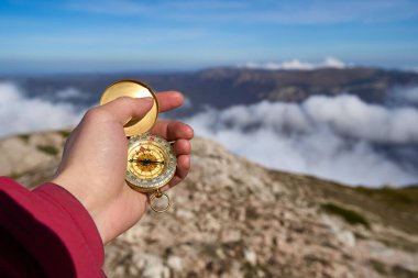 Closeup hand holding a compass with mountain and clouds background. Closeup of man tourist hand holding old magnetic compass on rock. Cloudy mountains on the background. Showing route. The four winds