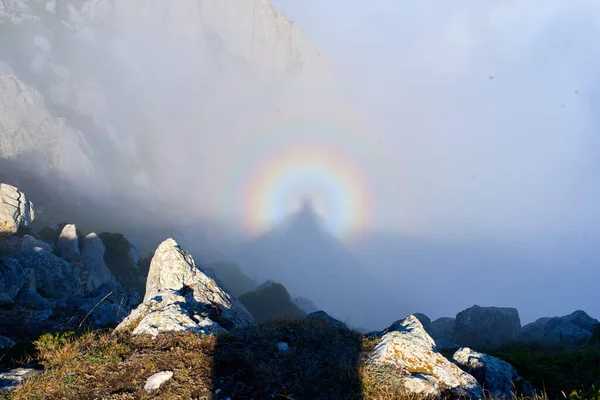 Rocky mountain crest in summer with fog and full rainbow in the background. Rocky hills in haze with round rainbow. Triple rainbow in clouds at sunrise. Rocky mountain top ridge on a foggy day