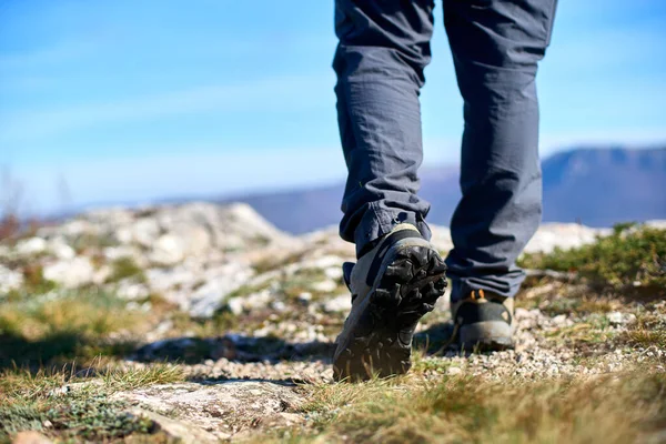 Legs of a hiker in trekking boots walking in mountains closeup shot. Feet of walking tourist wearing trekking shoes on a rocky road captured from behind. Hiking male wearing pants and boots walk