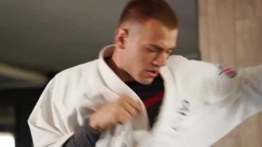 Martial arts man in kimonos practices kicks and punches. Male fighter wearing kimono training at gym. Kick boxer jumping and practicing kicking and punching actions indoors. People and sport concept