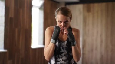 Kadın boksör tek başına spor salonunda kameraya bakarak yumruk hareketleri yapıyor. Elleri bandajlı, kameraya bakan ve zıplayan yumruklar atan motive olmuş kadın dövüşçü.
