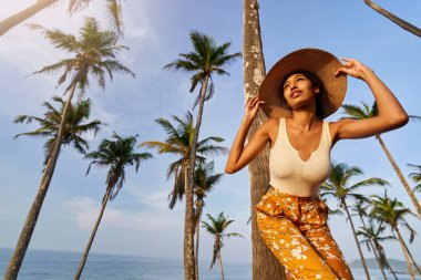 Young african female model posing in colorful clothes at tropical setting at sunrise. Black woman against exotic scenery at dawn. Multiracial dark-skinned model poses in front of palm trees at sunset