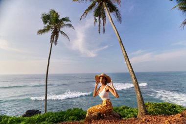 Young african female model posing in colorful clothes at scenic tropical location by ocean between palms at sunrise. Black woman sitting against exotic scenery with view of sea and palm trees at dawn