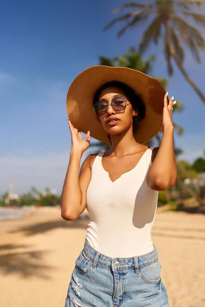 Young african female model in a straw hat and sunglasses posing at resort by sea at sunrise. Black woman against scenic rocky green island and ocean tide at dawn. Multiracial model poses on vacation
