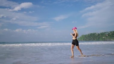 Young blonde woman jogs by sea at sunrise. Caucasian female runs at beach by ocean at dusk. Fit sporty girl exercises outdoor with tropical background. Healthy lifestyle, body. Side view tracking shot