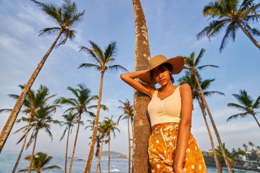 Young african female model posing in colorful clothes at tropical setting at sunrise. Black woman against exotic scenery at dawn. Multiracial dark-skinned model poses in front of palm trees at sunset