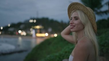 Young blonde woman in swimwear and straw hat on edge of picturesque hill with sea view and lights on background after sunset smiling enjoying the view alone. Caucasian young female in healing place