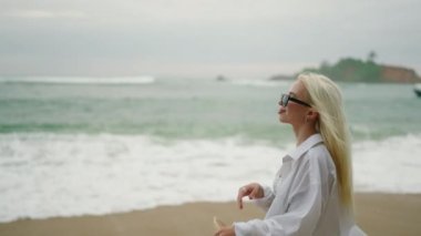 Slim blonde young woman in white shirt and onepiece swimsuit with straw hat wearing sunglasses walks along the beach closeup. Happy pretty female walking by sea portrait. Girl enjoys summer