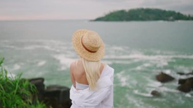 Young blonde woman sitting alone on bench self reflecting with spectacular view of ocean view from back. Blonde girl in white gown relaxes on bench at scenic tropical seaview location enjoying view