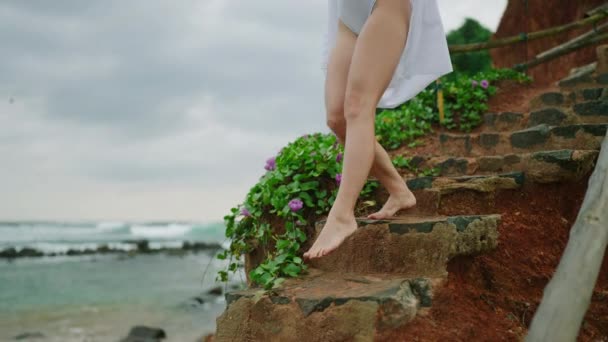 身穿泳衣和白衬衫的小女孩赤脚走过石山特写 雌性的腿和脚走下楼梯 被切割成红色的岩石 年轻女子乘天然石阶下山观景山 — 图库视频影像