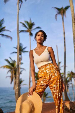 Young african female model posing in colorful clothes at tropical setting at sunrise. Black woman against exotic scenery at dawn. Multiracial dark-skinned model poses in front of palm trees at sunset
