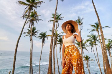 Young african female model posing in colorful clothes at tropical setting at sunrise. Black woman against exotic scenery at dawn. Multiracial dark-skinned model poses in front of palm trees at sunset