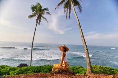 Young african female model posing in colorful clothes at scenic tropical location by ocean between palms at sunrise. Black woman sitting against exotic scenery with view of sea and palm trees at dawn