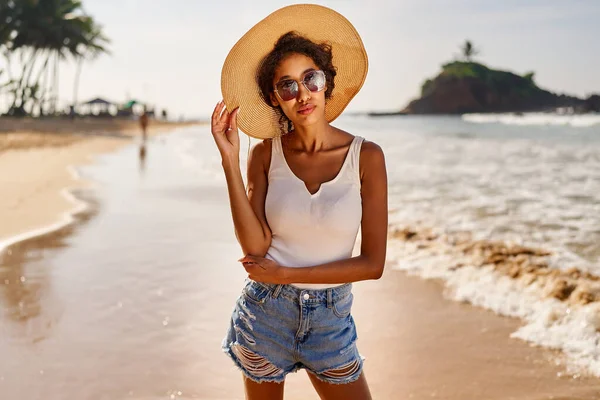 Young african female model in a straw hat and sunglasses posing at resort by sea at sunrise. Black woman against scenic rocky green island and ocean tide at dawn. Multiracial model poses on vacation