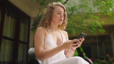 Young woman using smart phone texting with AI, surfing social media, checking news, playing mobile games and texting messages sitting under tree. Millennial lady spending time with cell gadget outside