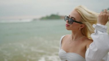 Slim blonde young woman in white shirt and onepiece swimsuit wearing sunglasses walks along beach touches hair looks at sea and smiles. Happy pretty caucasian female walking by sea enjoying herself